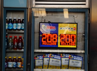 An advertisement display in a store window features lottery jackpot amounts for Mega Millions and Powerball, both showing jackpots of 208 million and 200 million respectively. Below the display, several lottery tickets and promotional posters are visible. To the left of the display, there is a refrigerator stocked with various drinks including different flavors of sports drinks and bottled water.