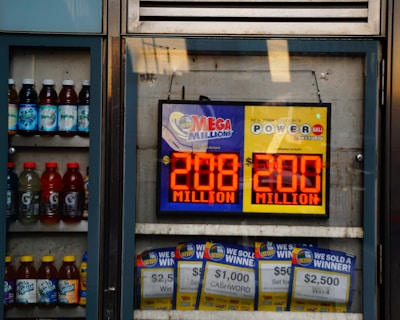 An advertisement display in a store window features lottery jackpot amounts for Mega Millions and Powerball, both showing jackpots of 208 million and 200 million respectively. Below the display, several lottery tickets and promotional posters are visible. To the left of the display, there is a refrigerator stocked with various drinks including different flavors of sports drinks and bottled water.