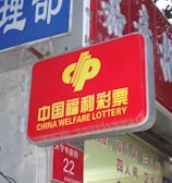 A red sign displaying the text 'China Welfare Lottery' in both Chinese and English, mounted on the exterior of a building. The surroundings include a blue sign with Chinese characters and a partial tree trunk visible on the right side.