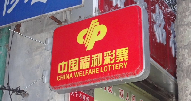 A red sign displaying the text 'China Welfare Lottery' in both Chinese and English, mounted on the exterior of a building. The surroundings include a blue sign with Chinese characters and a partial tree trunk visible on the right side.
