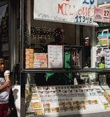 A child holding an ice cream cone stands outside a small kiosk or lottery ticket booth. The booth displays various colorful lottery tickets in the window. There are signs advertising a significant lottery prize amount in Spanish. A person can be seen partially obscured inside the booth, and plants are visible around the area, suggesting an outdoor setting.