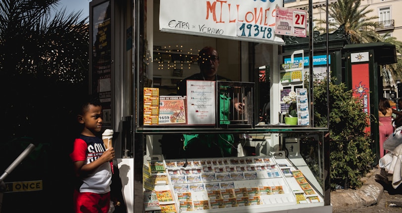 A child holding an ice cream cone stands outside a small kiosk or lottery ticket booth. The booth displays various colorful lottery tickets in the window. There are signs advertising a significant lottery prize amount in Spanish. A person can be seen partially obscured inside the booth, and plants are visible around the area, suggesting an outdoor setting.