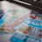 A selection of colorful lottery scratch-off tickets placed on a flat surface near a window. The image has a shallow depth of field, focusing on the tickets while blurring the background. The tickets display various games and prize amounts.