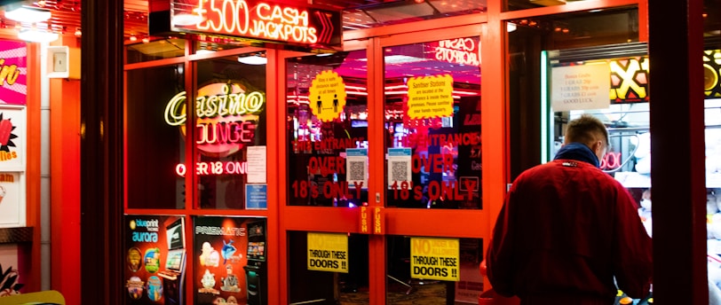 A brightly lit entrance to a casino with neon signs, including one advertising a £500 jackpot. The doors have clear signage indicating entry is restricted to those over 18. A person in a red jacket stands to the right, possibly interacting with a machine or display.