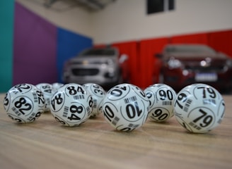 A collection of white lottery balls with black numerals scattered on a wooden surface. Two vehicles are blurred in the background, parked in a room with multicolored walls including teal, purple, blue, and red panels.