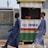 Two individuals are walking past a small kiosk with signage for the National Lottery Authority. The kiosk, decorated with a sign that reads 'Full Payment,' features yellow, green, red, and white colors. The individuals are dressed in matching long blue robes and hats, with one wearing black boots and the other white boots.
