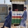 Two individuals are walking past a small kiosk with signage for the National Lottery Authority. The kiosk, decorated with a sign that reads 'Full Payment,' features yellow, green, red, and white colors. The individuals are dressed in matching long blue robes and hats, with one wearing black boots and the other white boots.