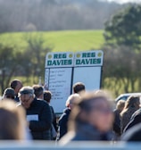 A group of people gathered outdoors with two large betting boards in the background labeled 'REG DAVIES'. The setting appears to be a countryside location with trees and fields visible in the distance. The crowd consists of individuals dressed in jackets and hats, engaging in conversation.