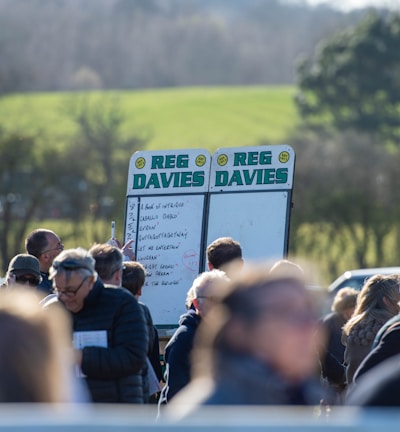 A group of people gathered outdoors with two large betting boards in the background labeled 'REG DAVIES'. The setting appears to be a countryside location with trees and fields visible in the distance. The crowd consists of individuals dressed in jackets and hats, engaging in conversation.