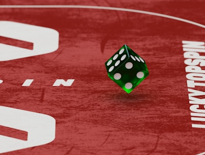 Two green dice with rounded edges are positioned on a red surface that seems to be part of a game board. The surface has white text and numbers on it, including the word 'LUCKYTOPSPIN' in capital letters.