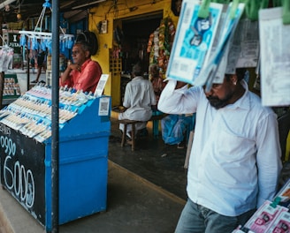 A street vendor stall displaying an array of lottery tickets clipped to a bright blue kiosk. A man in a red shirt leans on the counter, appearing thoughtful, while another man in a white shirt adjusts his hat and walks by. A variety of products hangs from strings above, and a colorful display fills the background with vibrant signs and packets. The scene suggests a busy, lively marketplace.