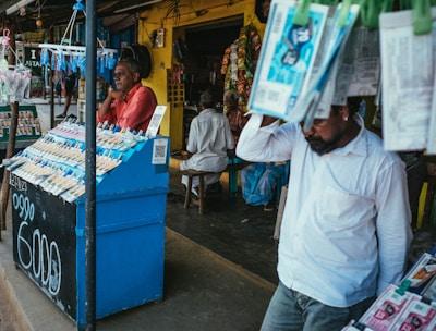 A street vendor stall displaying an array of lottery tickets clipped to a bright blue kiosk. A man in a red shirt leans on the counter, appearing thoughtful, while another man in a white shirt adjusts his hat and walks by. A variety of products hangs from strings above, and a colorful display fills the background with vibrant signs and packets. The scene suggests a busy, lively marketplace.